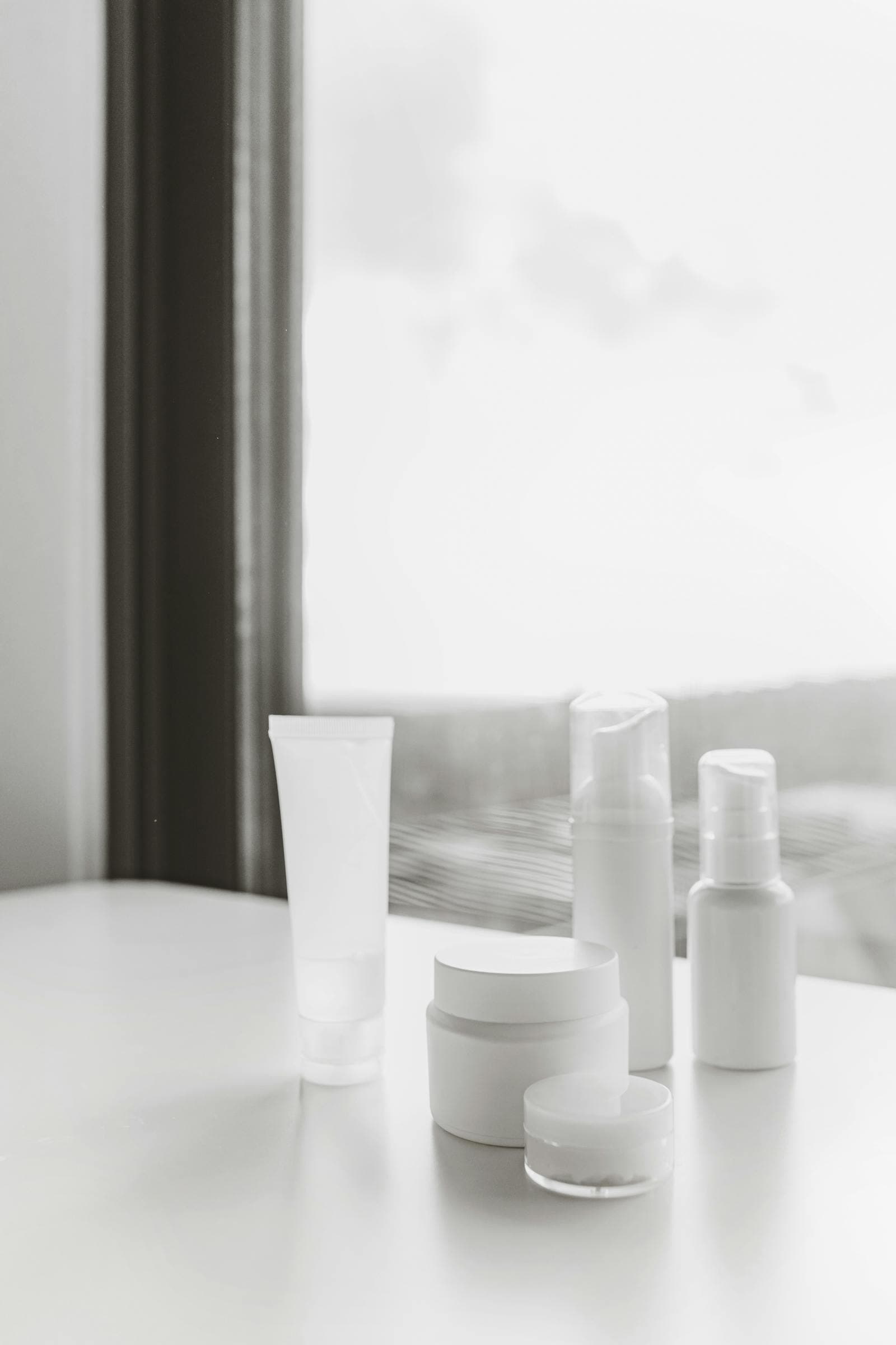 Minimal white skincare bottles and jars arranged on a table beside a bright window.